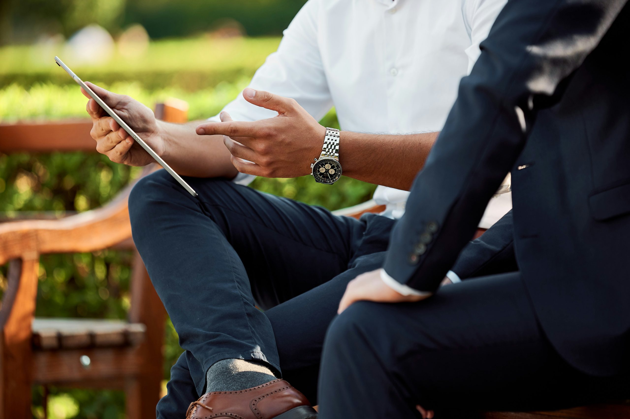 Due uomini in abiti da lavoro siedono all'aperto su una panchina. Uno dei due tiene un tablet e gesticola mentre parla, suggerendo una discussione o una riunione. Lo sfondo è una vegetazione sfocata. Sono visibili solo il busto e le mani.