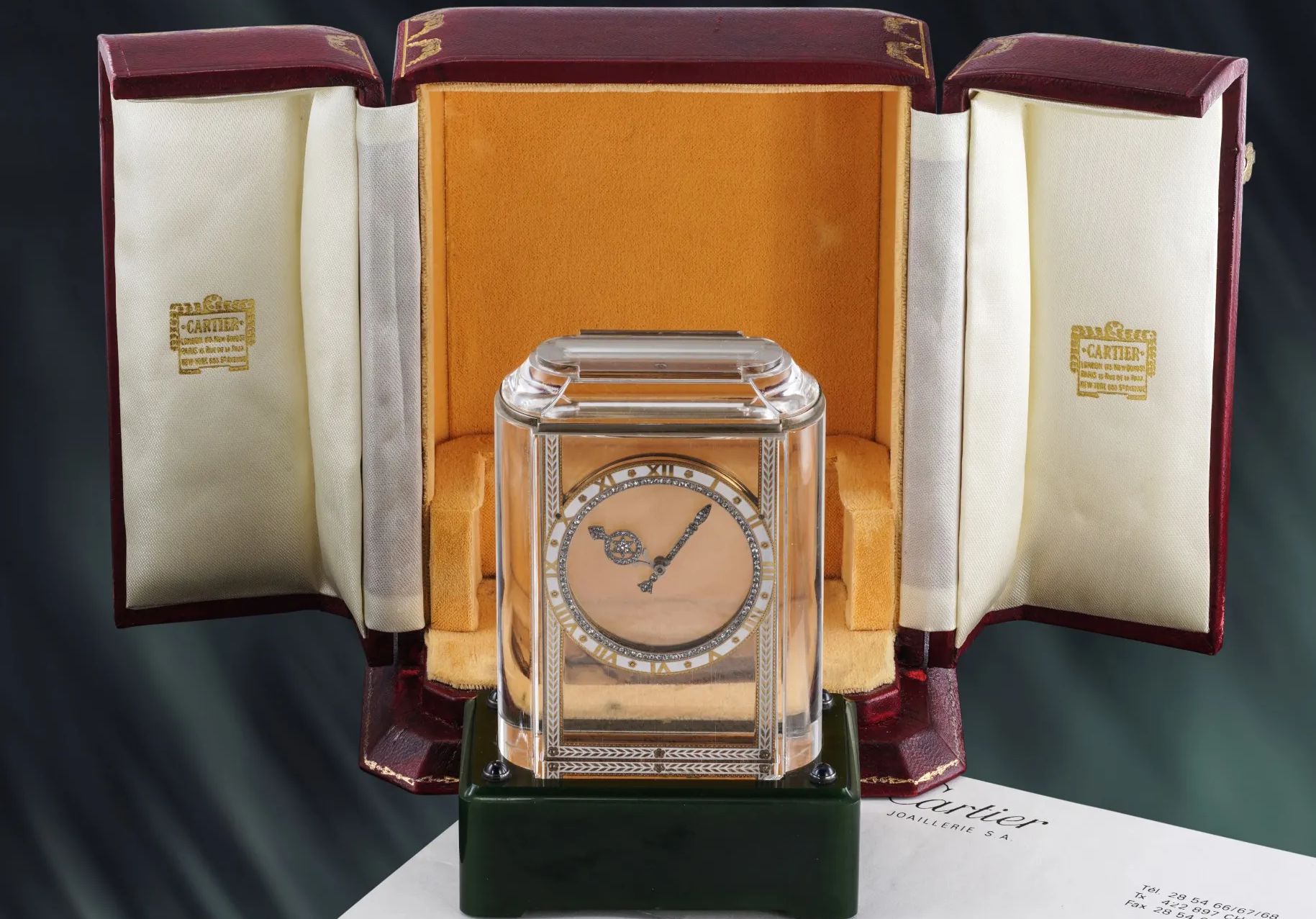 Un orologio Cartier con cassa trasparente e quadrante d'oro dettagliato si trova in una scatola di presentazione aperta di colore rosso e crema, rivestita di scritte in oro che ricordano i tesori dell'asta di orologi JP Morgan. Un documento con la scritta "Cartier" è parzialmente visibile in primo piano.