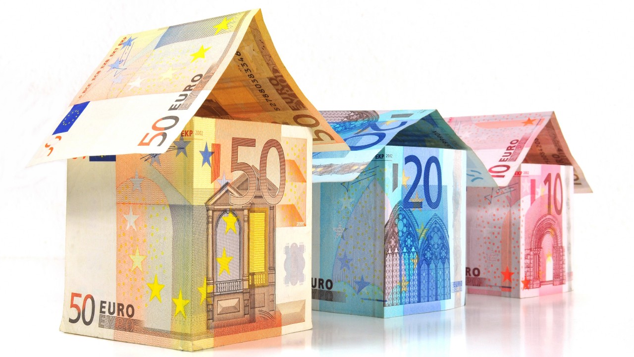 Three small house shapes are made from folded euro banknotes, including 50, 20, and 10 euro notes, arranged in a row on a white background.
