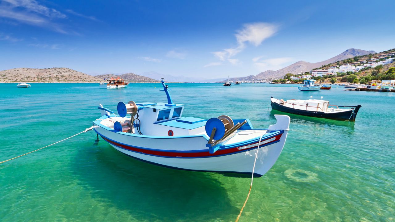 A white and blue fishing boat is anchored on clear turquoise water with mountains and a small coastal town in the background under a bright blue sky. Other boats are visible in the distance.