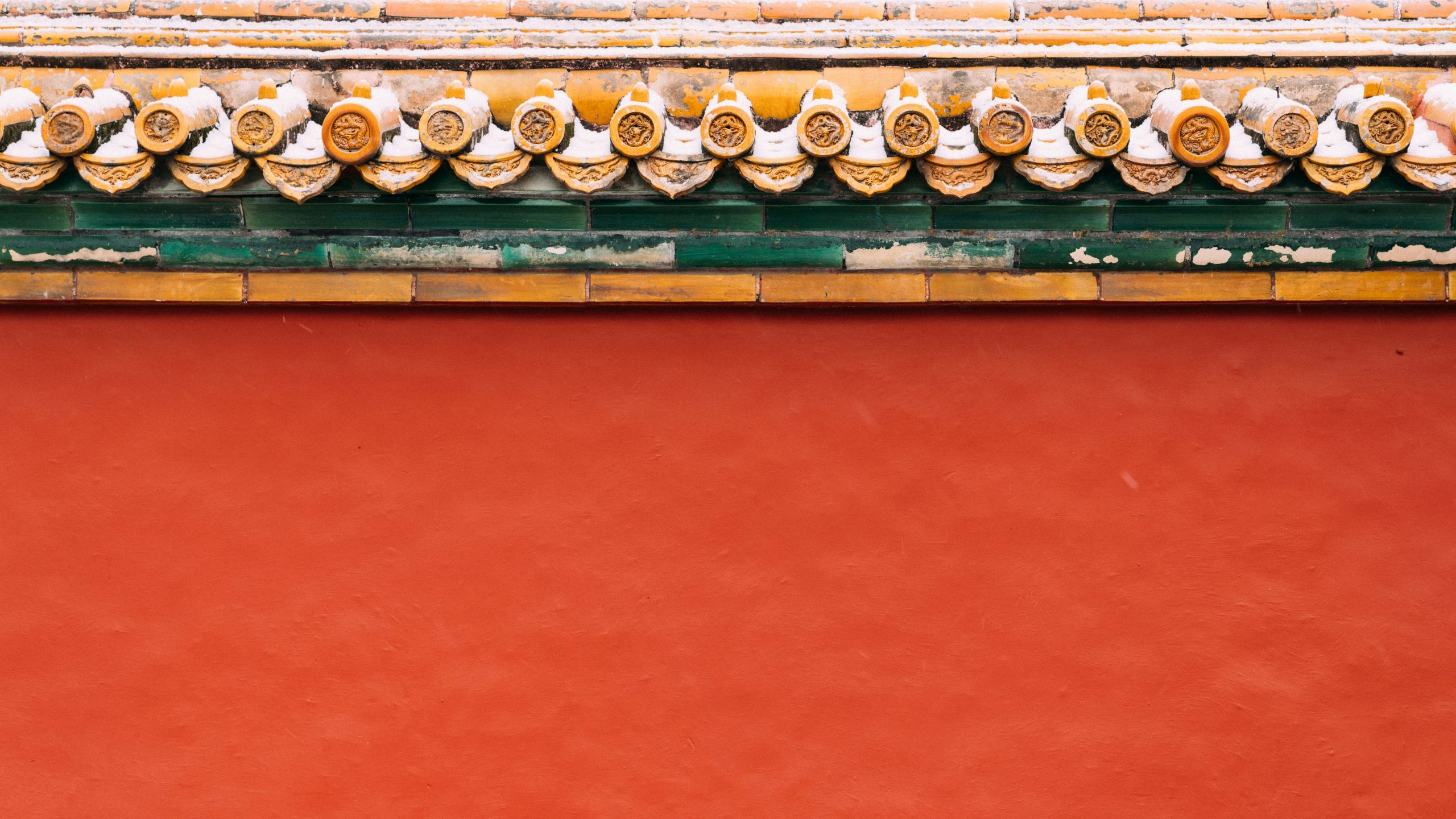 Primo piano di un tetto tradizionale cinese con tegole gialle decorate e un muro rosso sottostante, con finiture verdi e bianche. Le tegole sono ricoperte di neve e creano un contrasto con i colori vivaci.