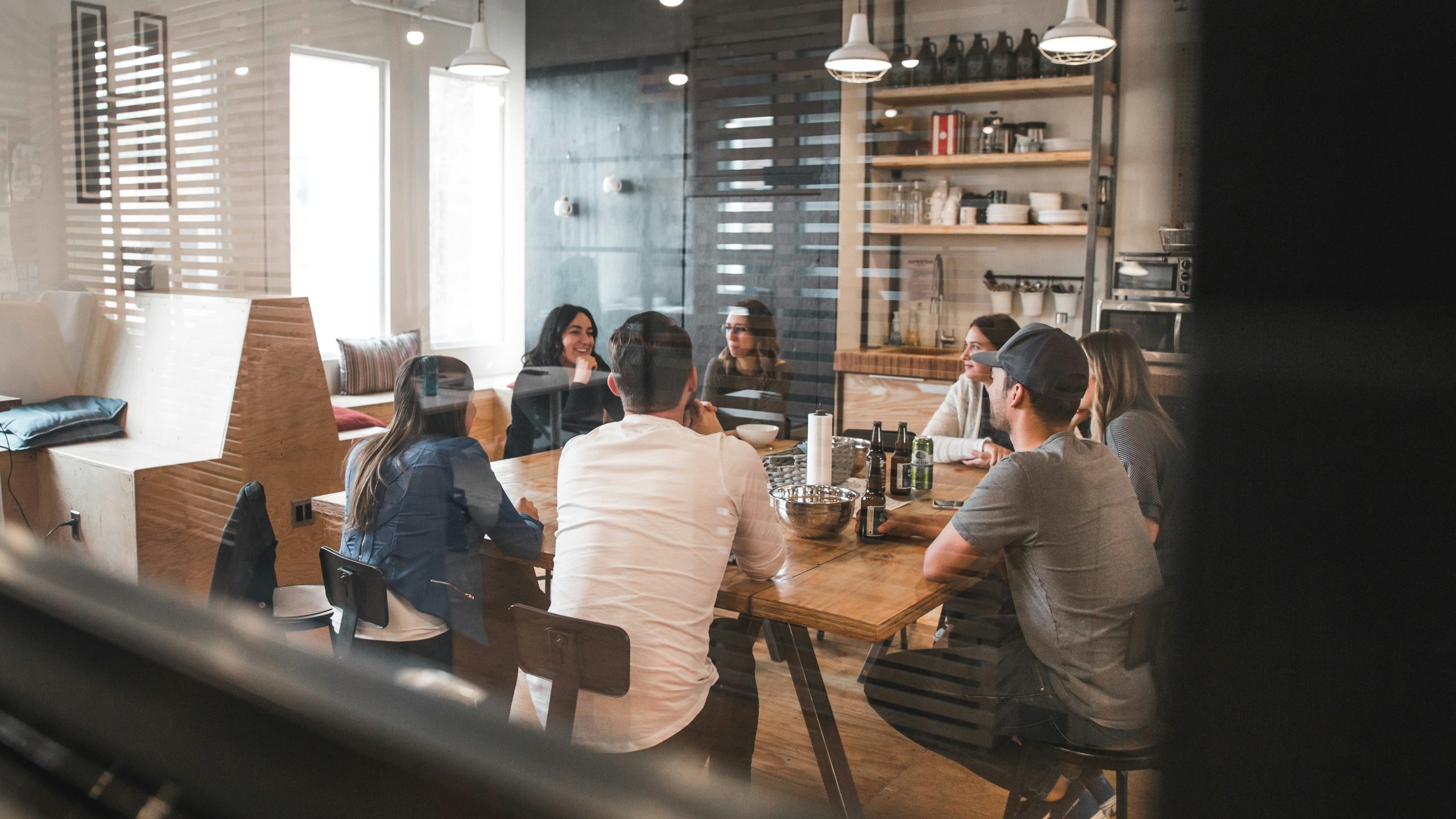 A group of people sit around a wooden table in a modern office, engaged in a meeting. Shelves with dishes and bottles are visible in the background, and natural light comes in through large windows.