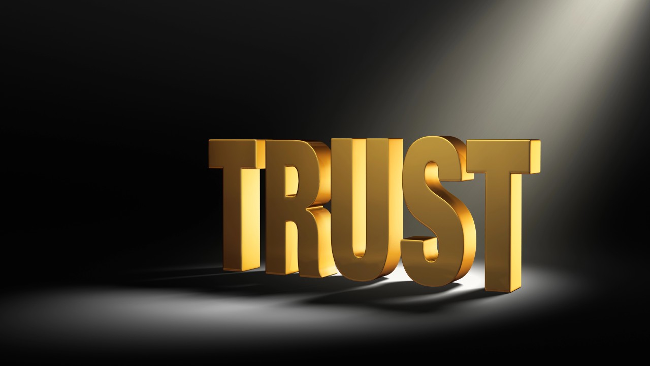 The word "TRUST" is displayed in large, bold, gold 3D letters under a spotlight, casting shadows on a dark background.