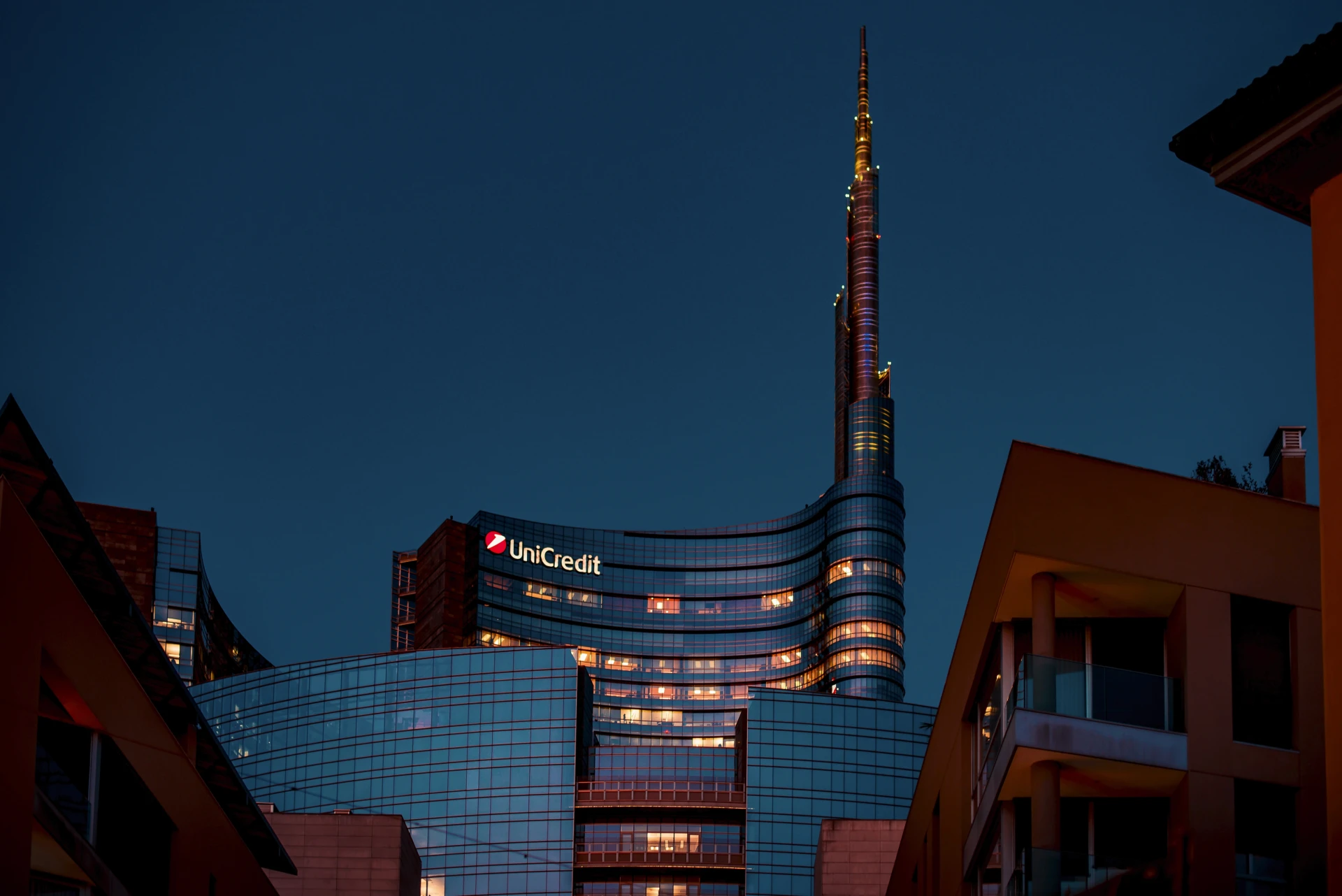 Un moderno grattacielo di vetro, l'UniCredit Tower di Milano, è illuminato al tramonto. Sede di importanti eventi finanziari come unicredit emissione obbligazioni, la sua alta guglia e le finestre illuminate si stagliano contro il cielo blu scuro della sera.