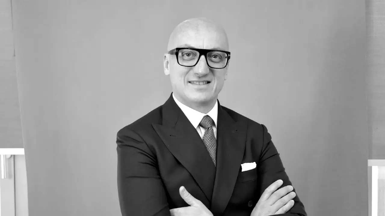Un uomo calvo che indossa occhiali e un abito con cravatta si staglia su uno sfondo semplice, sorridendo con le braccia incrociate. L'immagine è in bianco e nero.