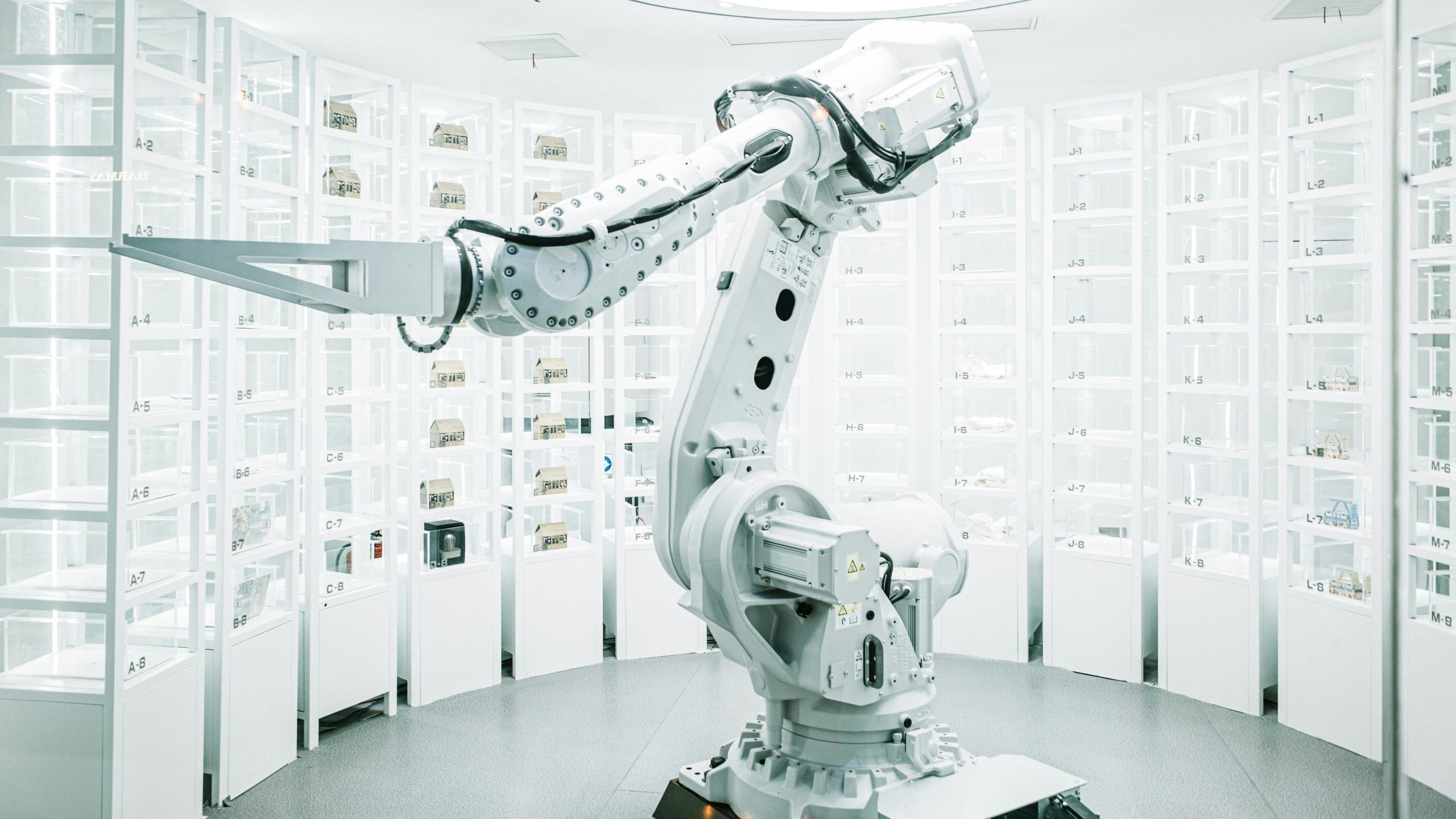 Un braccio robotico industriale bianco è posizionato al centro di una stanza luminosa e moderna, fiancheggiata da scaffali pieni di piccoli contenitori, che suggeriscono un ambiente di stoccaggio o di laboratorio automatizzato.