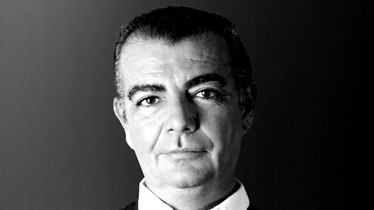 Ritratto in bianco e nero di un uomo di mezza età con capelli corti e scuri, che indossa un abito e una camicia bianca con colletto e guarda direttamente verso l'obiettivo con un'espressione neutra.