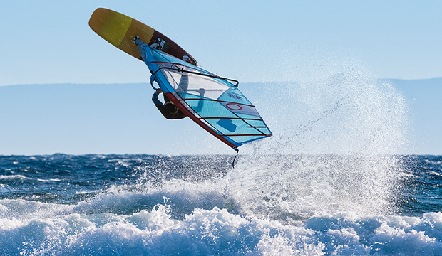 Un windsurfer esegue un salto sopra le onde dell'oceano, con schizzi d'acqua intorno alla tavola e alla vela sotto un cielo azzurro e limpido.