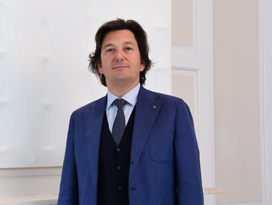 A man with medium-length dark hair wearing a blue suit, white shirt, and dark tie stands in a bright, modern room with white walls.