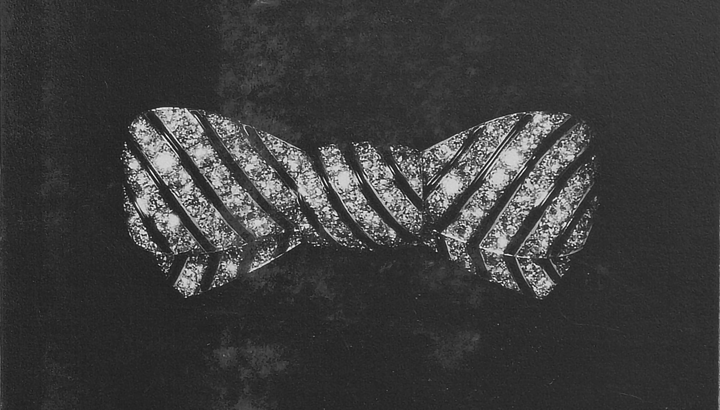 Una spilla tempestata di diamanti a forma di papillon si staglia su uno sfondo scuro e strutturato. La spilla presenta strisce diagonali di diamanti scintillanti, creando un aspetto elegante e lussuoso.