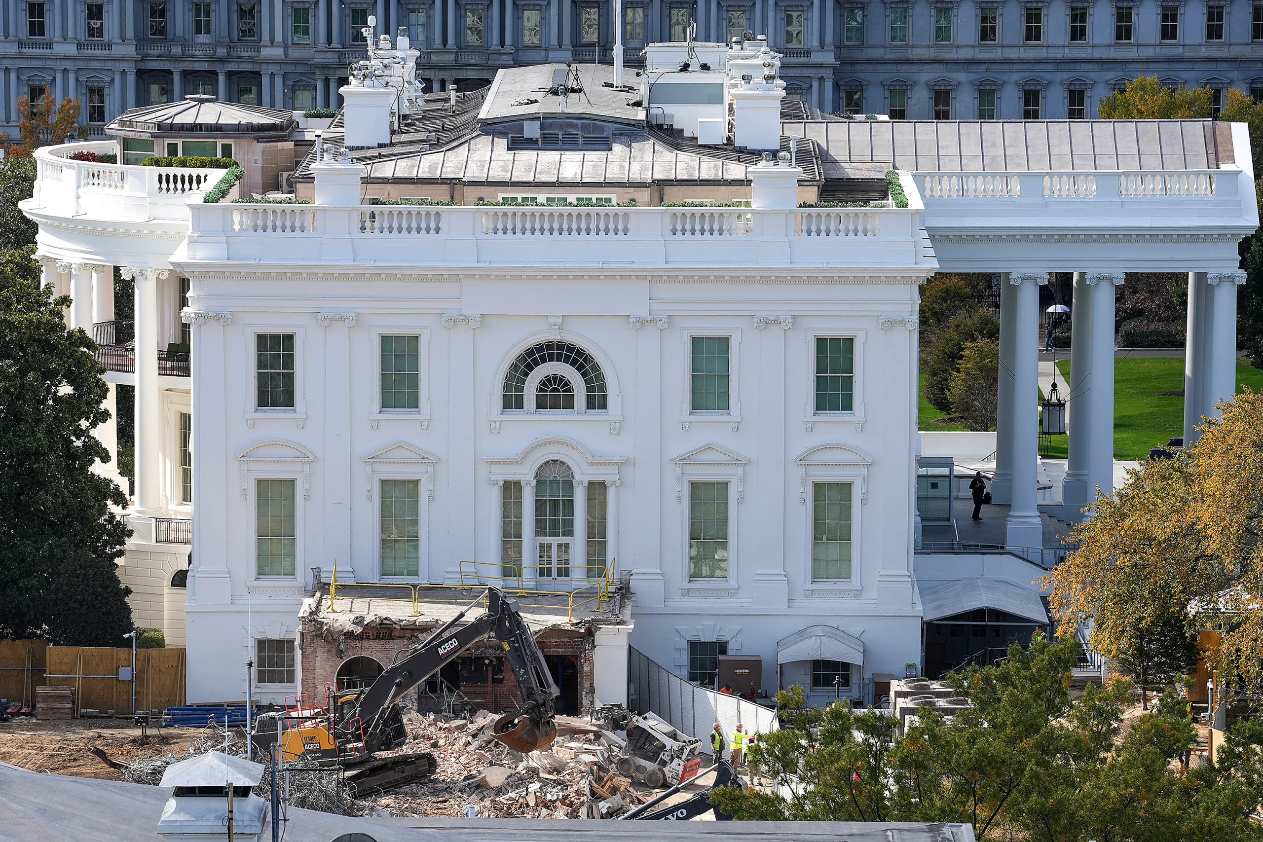 Un escavatore demolisce parte dell'esterno della Casa Bianca, con macerie sparse davanti e operai in tenuta di sicurezza nelle vicinanze. L'edificio retrostante mostra le classiche colonne bianche e le grandi finestre.