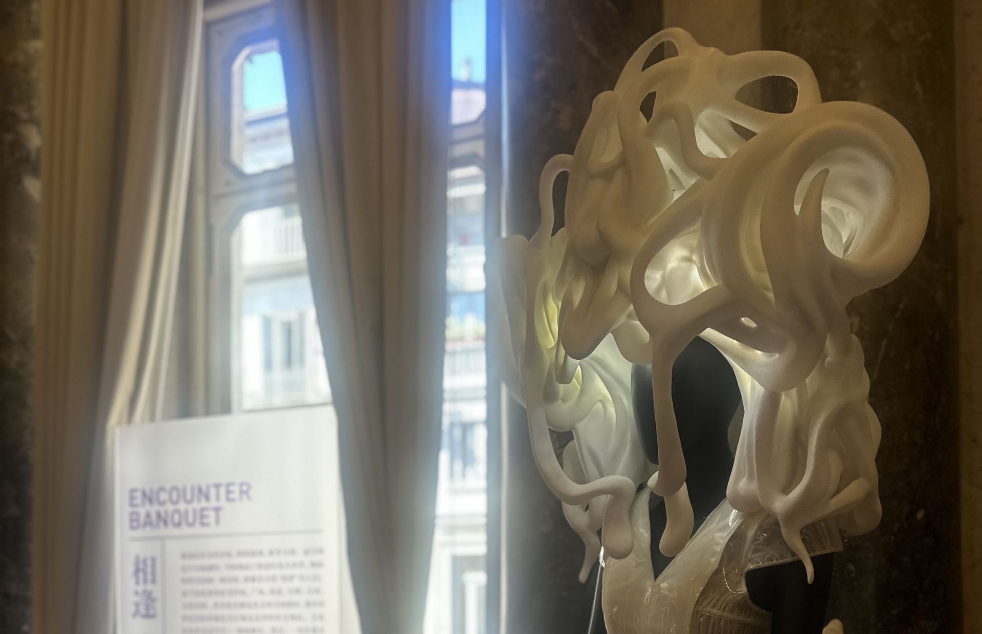 Un'opera d'arte scultorea bianca di forma organica è esposta all'interno, vicino ad alte finestre con tende drappeggiate. La luce del sole filtra e sullo sfondo è parzialmente visibile l'insegna "ENCOUNTER BANQUET".