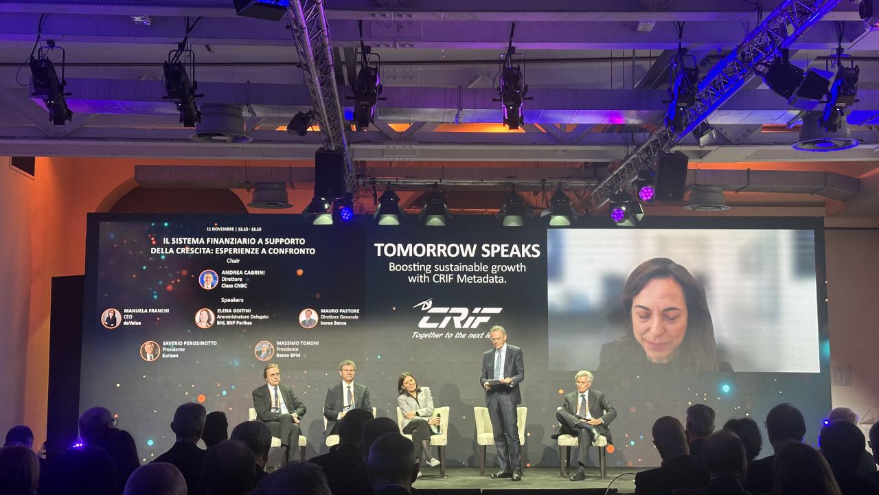 Un panel di conferenze sul palco con cinque relatori seduti e uno in piedi, che si rivolgono al pubblico. Un grande schermo alle loro spalle mostra "Tomorrow Speaks" e un partecipante alla videochiamata, con i dettagli dell'evento e i nomi dei relatori.