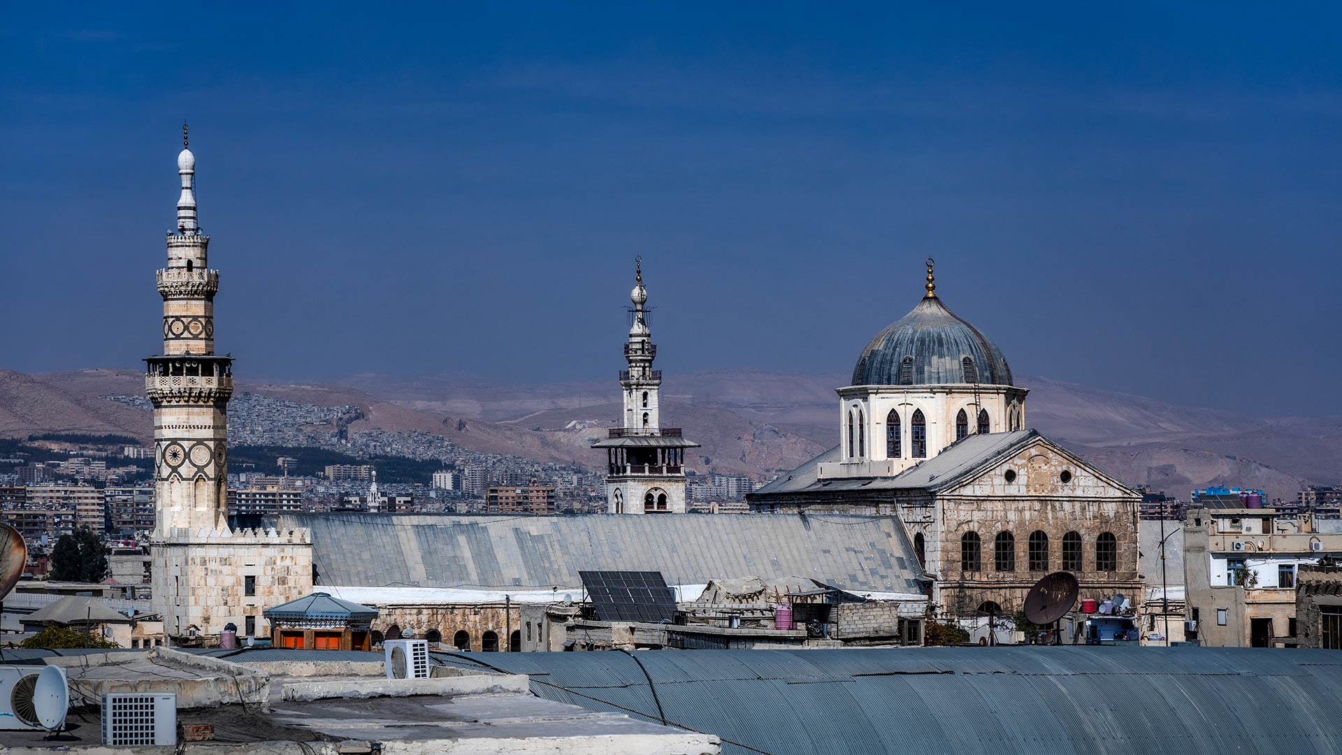 Una moschea storica con due alti minareti e un grande tetto a cupola si staglia sullo sfondo di edifici cittadini e montagne lontane, sotto un cielo azzurro e limpido.