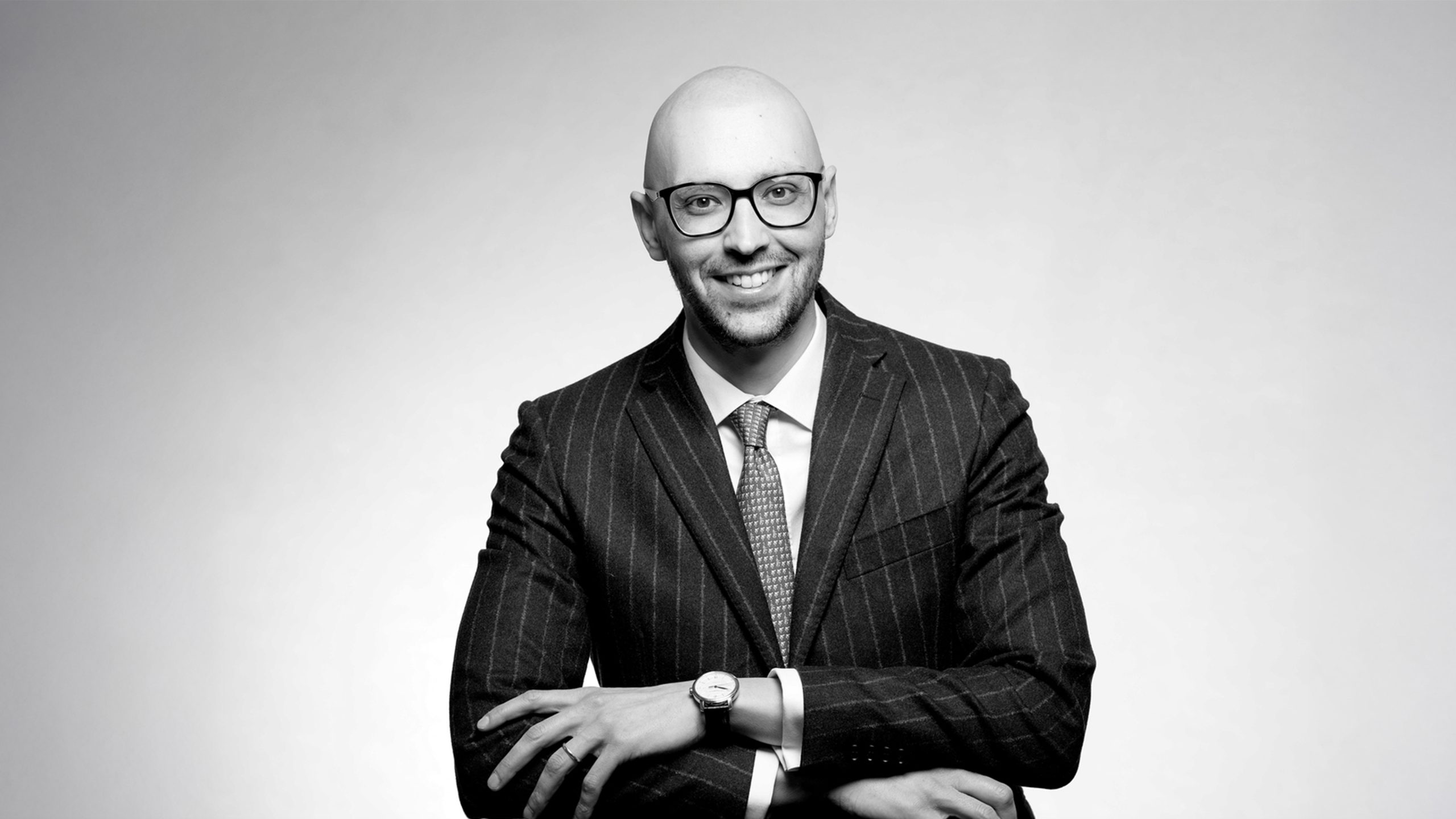Un uomo calvo con occhiali, abito gessato e cravatta siede con le braccia incrociate, sorridendo alla macchina fotografica su uno sfondo chiaro e semplice, che trasmette fiducia nella pianificazione patrimoniale e nel patrimonio. La foto è in bianco e nero.