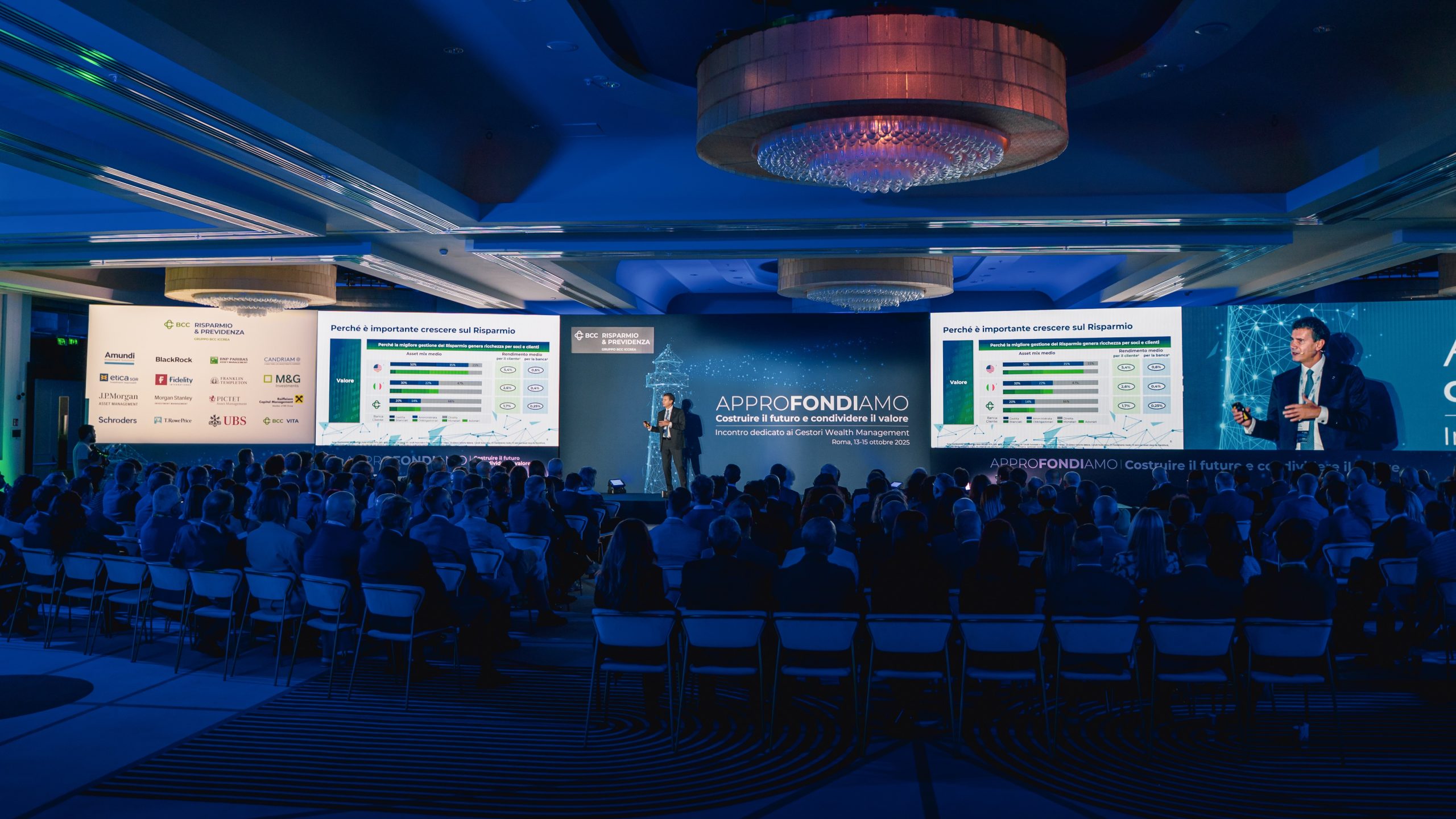 Un relatore presenta sul palco a un pubblico seduto a una conferenza, con grandi schermi che visualizzano grafici, diagrammi e loghi dei partner in una sala moderna e illuminata di blu.
