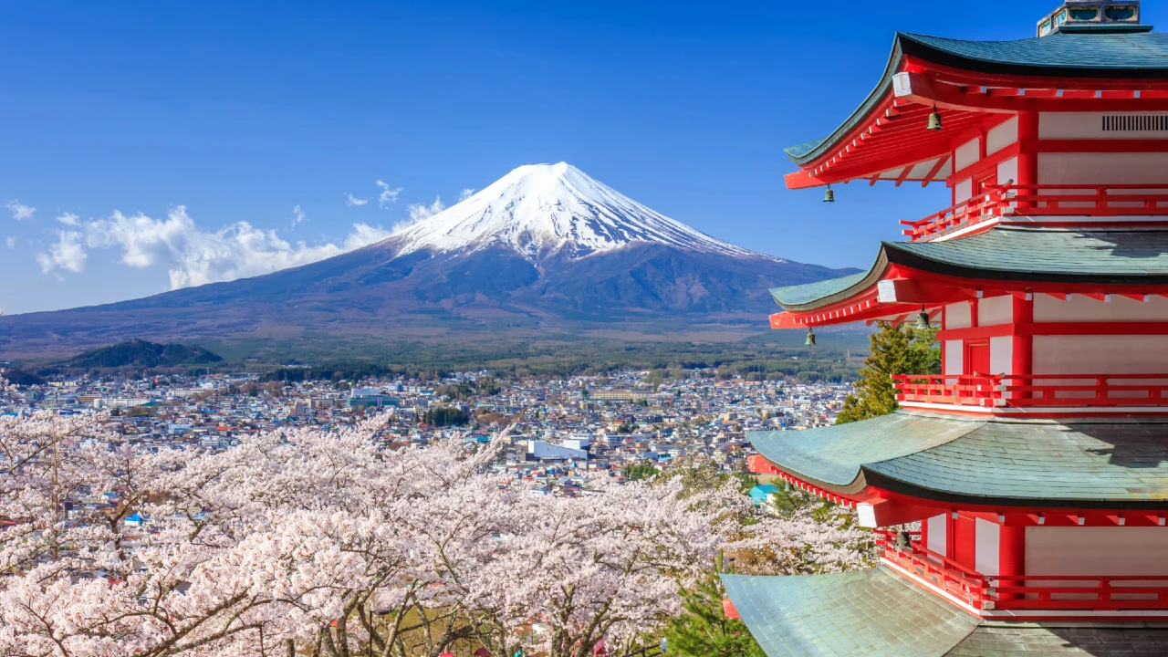 Una pagoda tradizionale giapponese si affaccia su alberi di ciliegio in fiore, con la cima innevata del Monte Fuji che si erge maestosa sullo sfondo sotto un cielo azzurro e limpido.