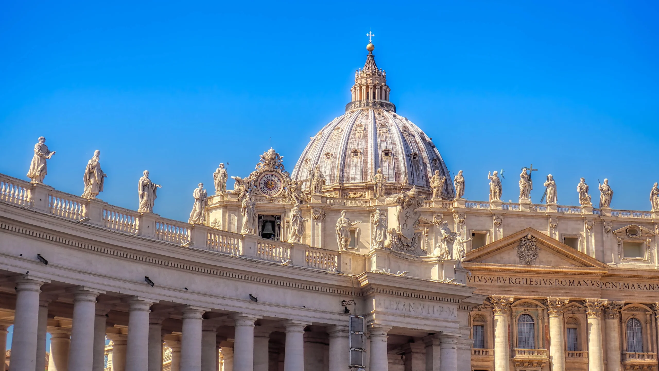 La Basilica di San Pietro nella Città del Vaticano con la sua grande cupola, le statue ornate, le colonne e gli intricati dettagli architettonici in un cielo azzurro e limpido.
