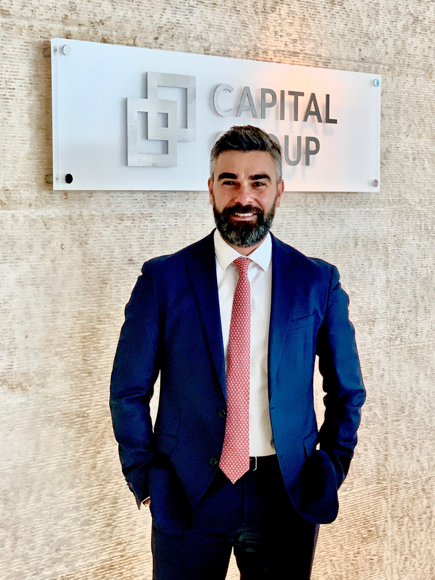 Un uomo in abito blu e cravatta rossa si trova sorridente davanti a un'insegna a muro con la scritta "CAPITAL GROUP" e un logo geometrico.