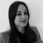 Ritratto in bianco e nero di una giovane donna con lunghi capelli scuri, che indossa un blazer su un top scuro, sorride leggermente e guarda l'obiettivo, su uno sfondo chiaro.