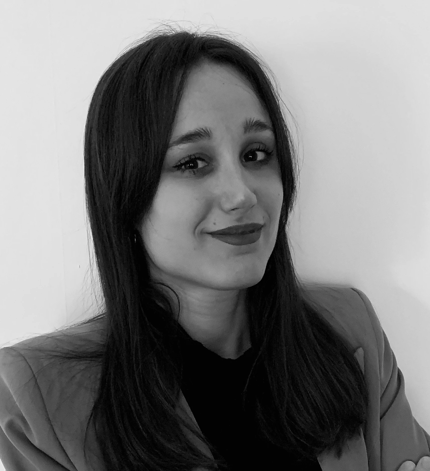 Ritratto in bianco e nero di una giovane donna con lunghi capelli scuri, che indossa un blazer su un top scuro, sorride leggermente e guarda l'obiettivo, su uno sfondo chiaro.