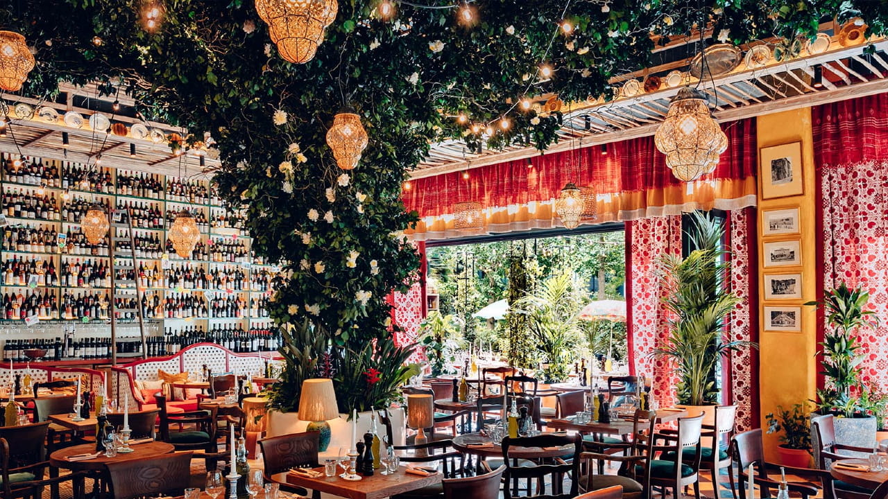 A vibrant restaurant interior with lush greenery, hanging lights, shelves filled with wine bottles, red patterned walls, and large windows opening to a garden. Wooden tables and chairs are neatly arranged.