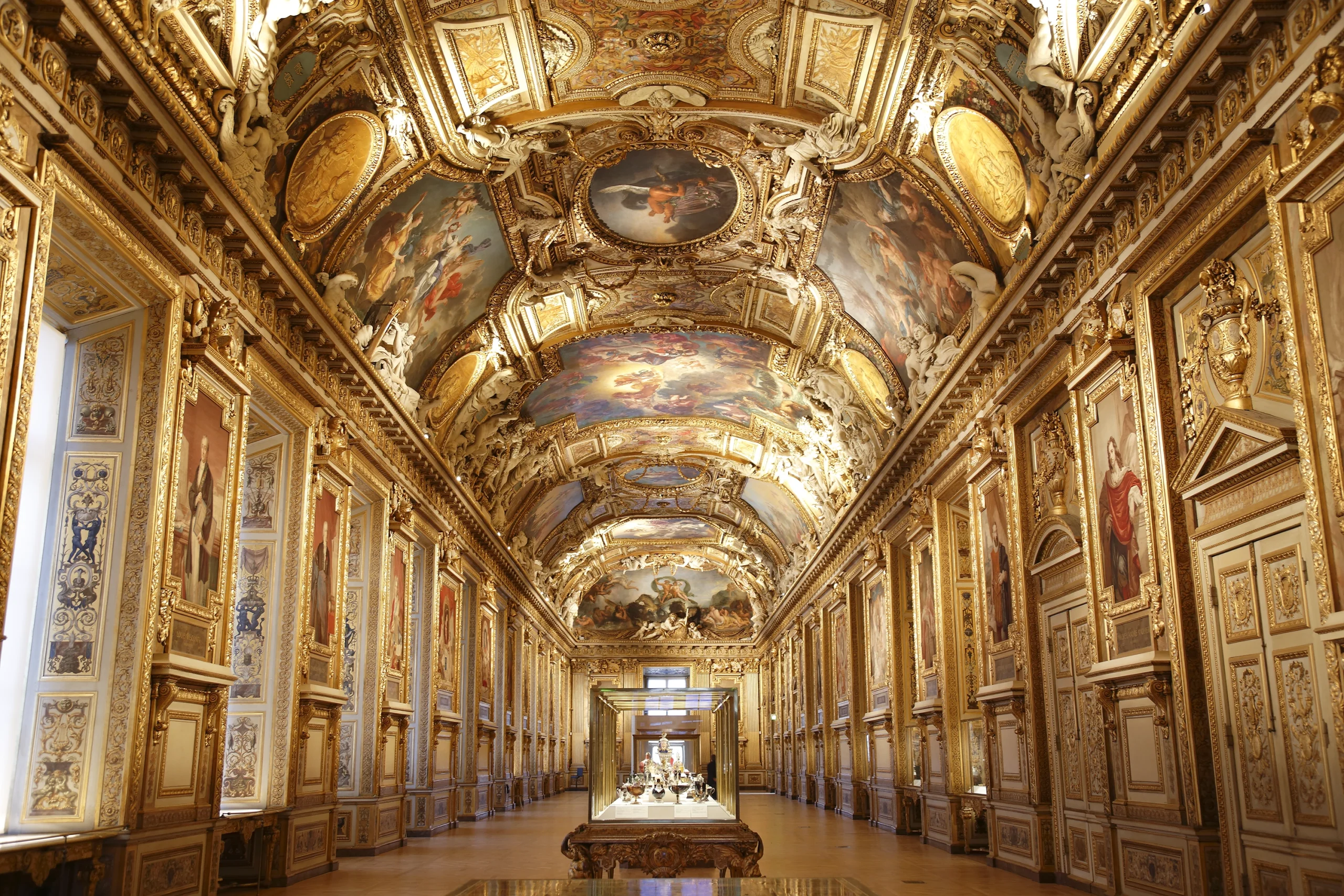 Una grande sala decorata con un elaborato soffitto a volta con dipinti dettagliati e accenti dorati, fiancheggiata da alte finestre e opere d'arte incorniciate, con una vetrina centrale in fondo.