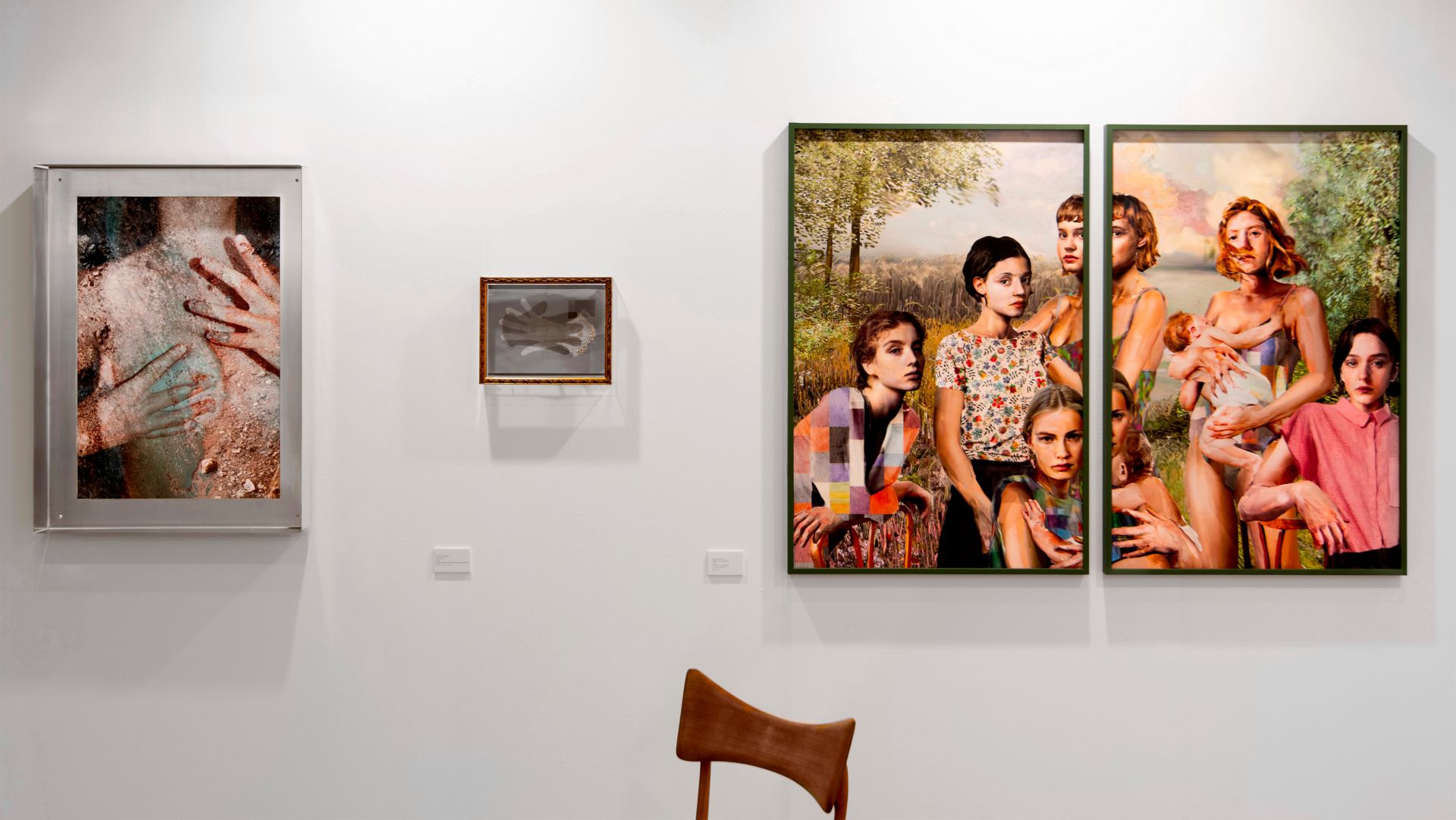 Una parete della galleria espone tre opere d'arte: una foto astratta di mani sulla pelle, un piccolo disegno incorniciato di una mano e un grande dipinto vivace di diverse donne all'aperto. Una sedia di legno si trova di fronte.