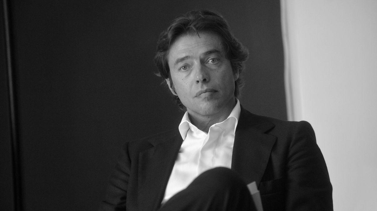 Un uomo in abito scuro e camicia bianca siede con un'espressione seria, guardando la macchina fotografica. La foto in bianco e nero, con uno sfondo semplice e bicolore, cattura l'essenza del ritratto di banca profilo.