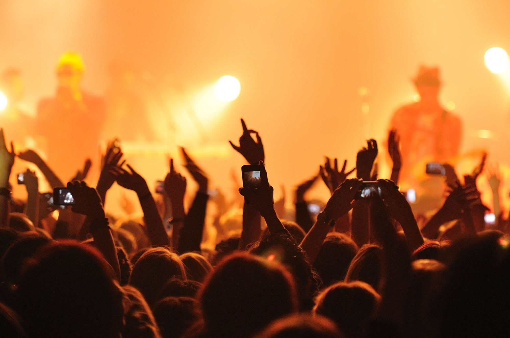 La folla di un concerto alza le mani e tiene in mano gli smartphone per registrare l'esibizione. Il palco è illuminato da luci arancioni e i musicisti sono visibili sullo sfondo.