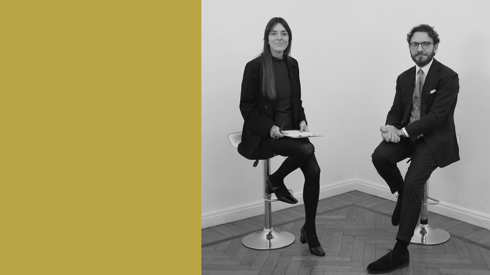 Una donna e un uomo in abiti da lavoro siedono su sgabelli da bar in una stanza minimalista con pavimenti in legno e una parete bianca. Il lato sinistro dell'immagine è riempito da un solido blocco giallo senape.