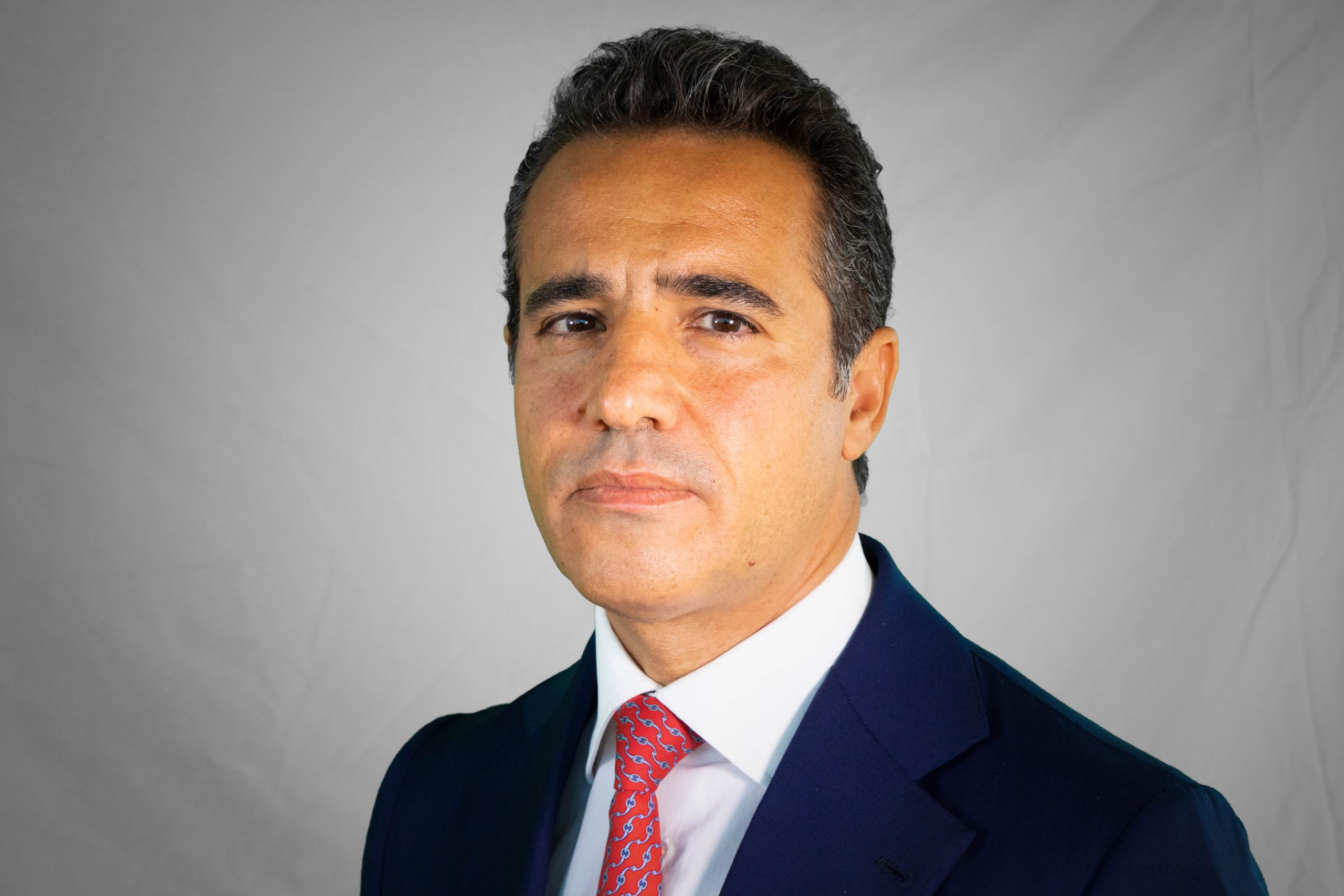 Un uomo con i capelli corti e scuri che indossa un abito blu navy, una camicia bianca e una cravatta a motivi rossi posa su uno sfondo grigio chiaro. È rivolto leggermente a sinistra e guarda seriamente la telecamera, riflettendo la professionalità nella gestione patrimoniale offerta da UniCredit.