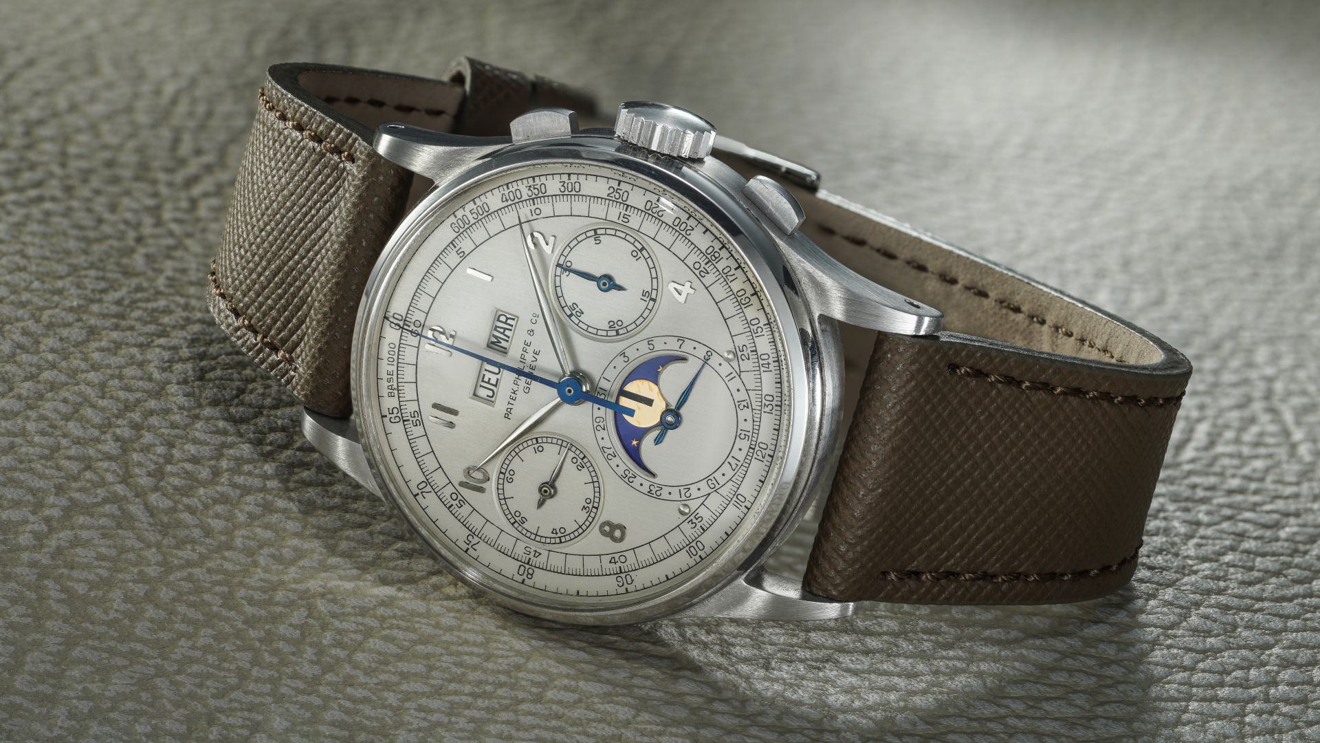 Orologio da polso di lusso color argento con cinturino in pelle marrone testurizzata, dotato di contatori multipli, finestrelle per il giorno e la data e indicatore delle fasi lunari, che poggiano su una superficie leggermente testurizzata.