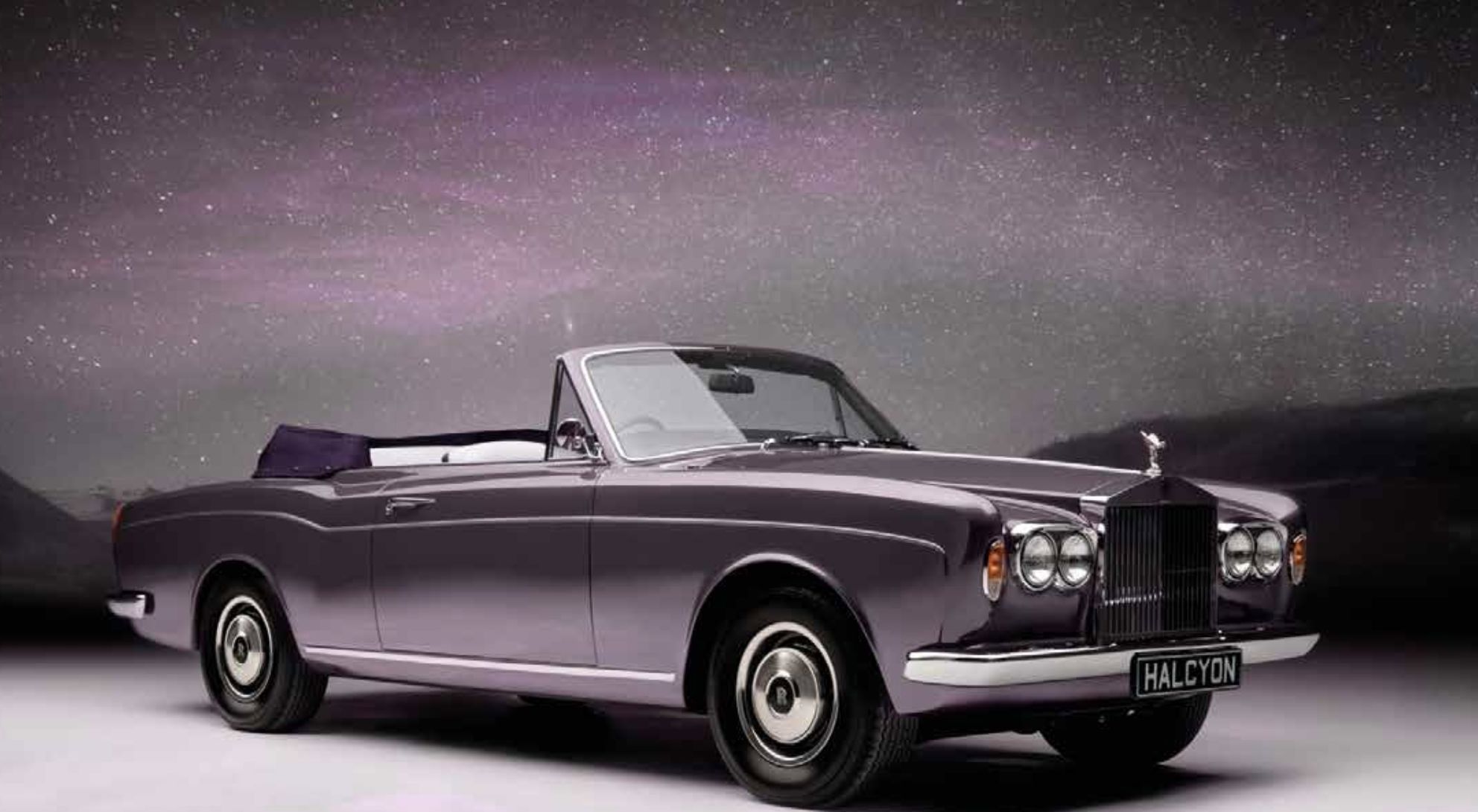 Una Rolls-Royce decappottabile viola d'epoca con la capote abbassata è parcheggiata sotto un cielo stellato, con "HALCYON" sulla targa.
