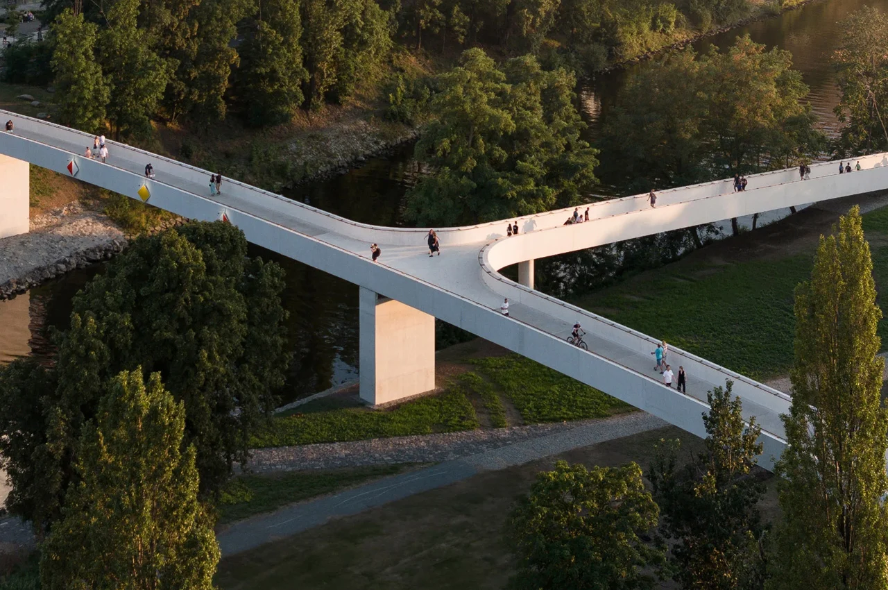 Un moderno ponte pedonale bianco con un design unico a forma di Y sovrasta un fiume, circondato da alberi verdi. Le persone camminano, vanno in bicicletta e si rilassano, proprio come gli investitori di private equity che cercano nuove prospettive da punti di osservazione elevati.