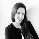 Una donna con lunghi capelli scuri sorride all'obiettivo. Indossa un blazer scuro su un top chiaro ed è in posa su un semplice sfondo bianco. L'immagine è in bianco e nero.
