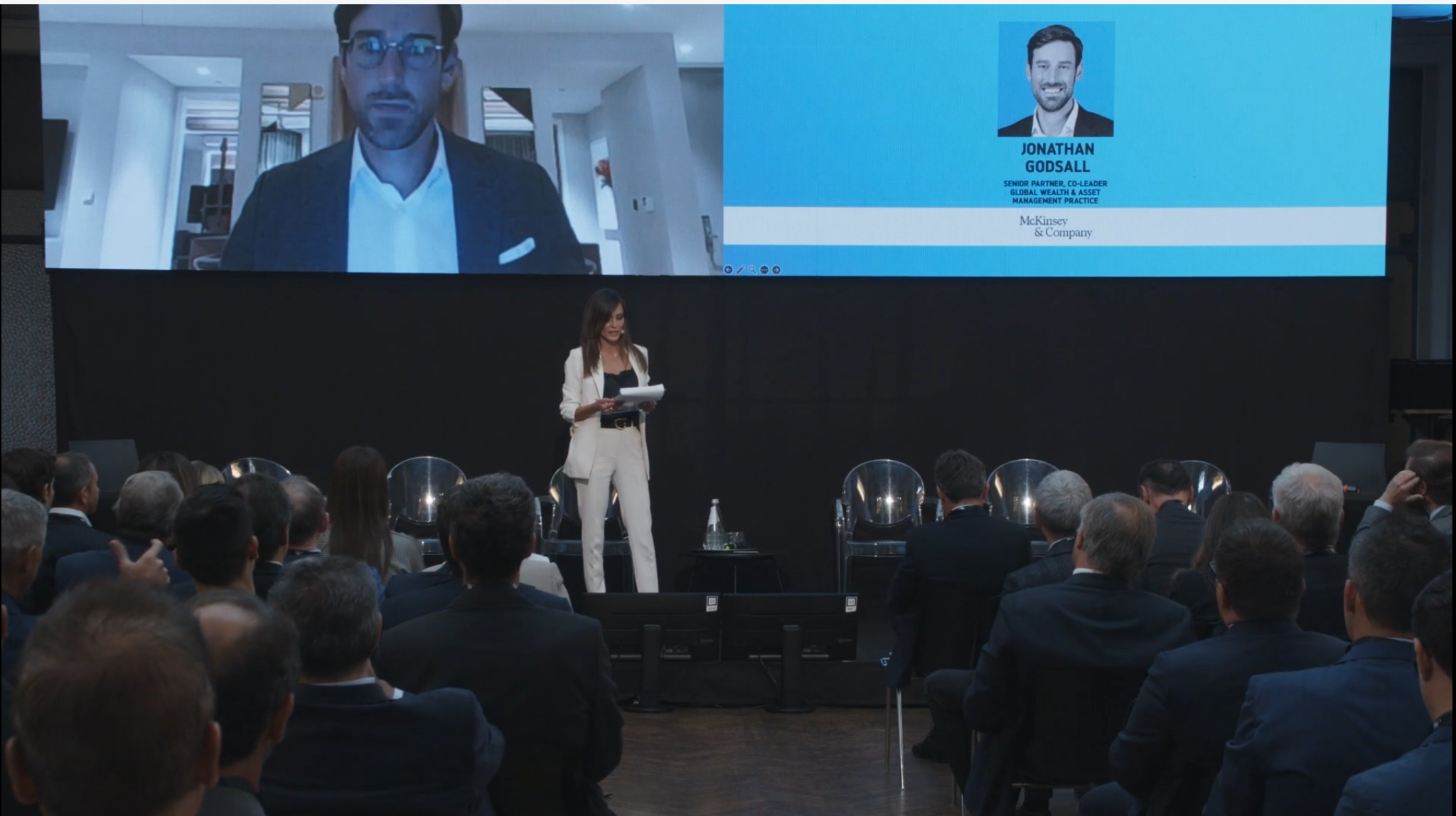 Una donna in abito bianco parla sul palco a un pubblico, con sedie vuote alle sue spalle. Un grande schermo mostra un uomo in giacca e cravatta in videochiamata e le informazioni del suo profilo, con la dicitura "Jonathan Gossall".