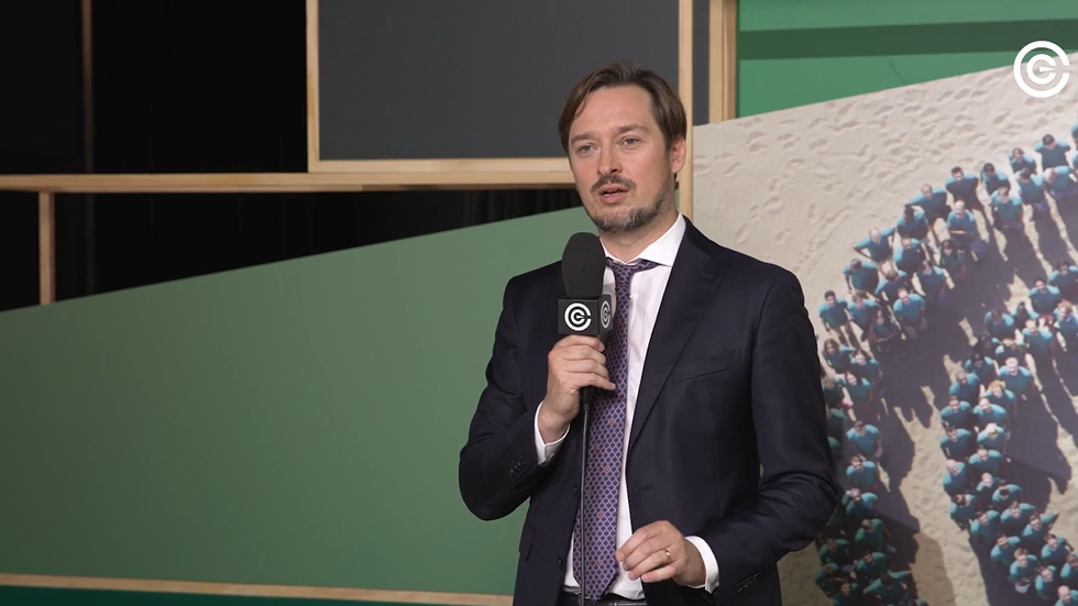 Un uomo in giacca e cravatta tiene un microfono mentre parla davanti a uno sfondo geometrico verde e beige con un logo circolare in alto a destra.