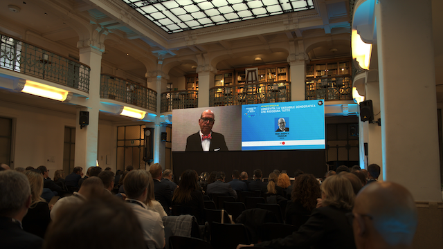 Un pubblico siede in una grande sala elegante con soffitti alti e librerie, guardando un oratore formale su uno schermo di proiezione durante una conferenza o un evento.