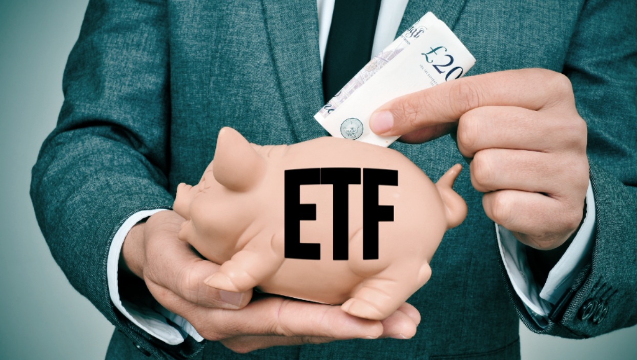 Una persona in giacca e cravatta mette una banconota da 20 sterline in un salvadanaio con l'etichetta "ETF", che simboleggia l'investimento o il risparmio in fondi negoziati in borsa.