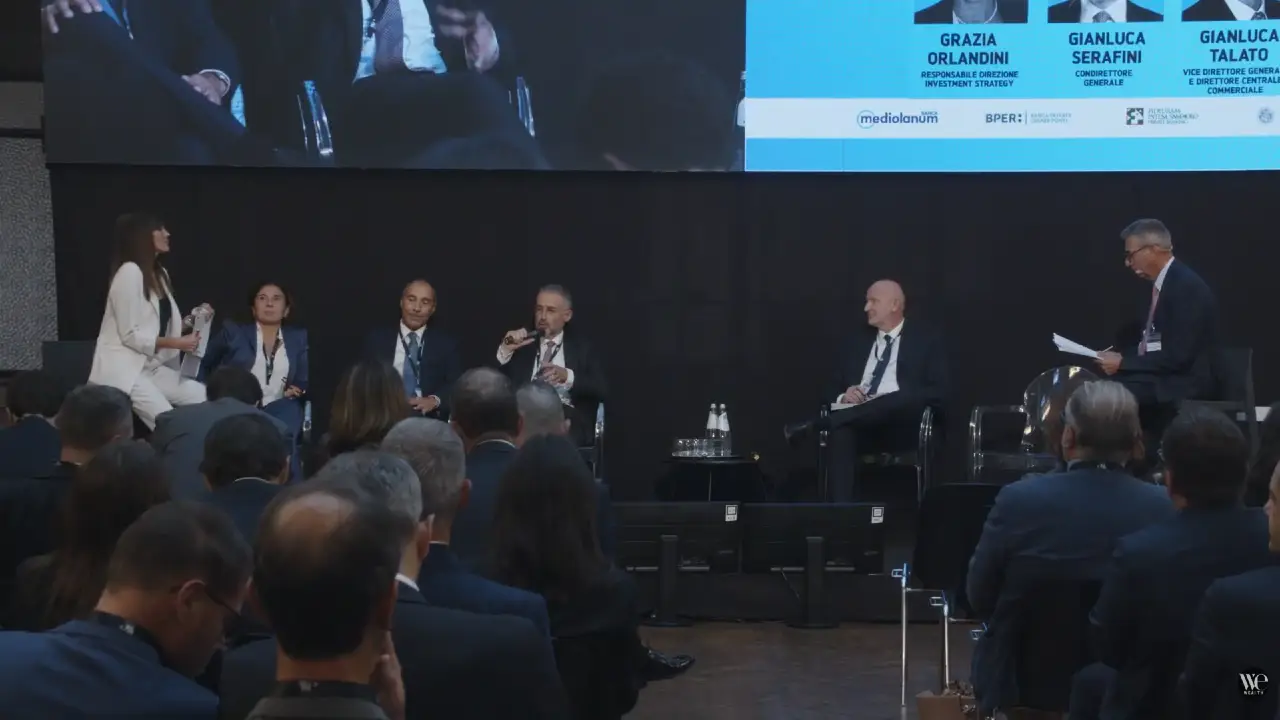Un gruppo di cinque persone in abiti da lavoro siede sul palco per discutere, mentre il pubblico ascolta. Una donna si alza e parla a sinistra. Uno schermo in alto mostra i nomi e i ruoli dei partecipanti.
