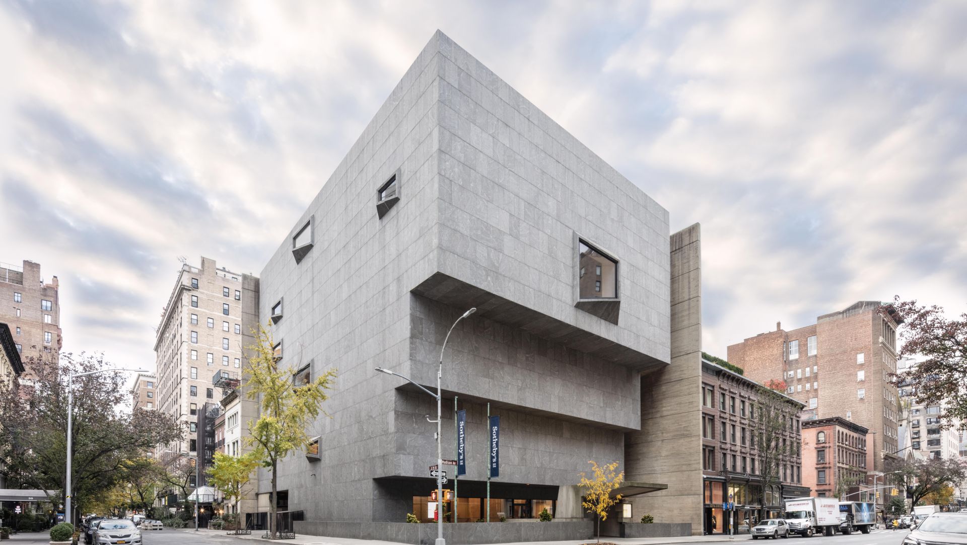 Un grande e moderno edificio in cemento con forme geometriche e angolari e piccole finestre, situato all'angolo di una strada cittadina e circondato da vecchi edifici in mattoni e alberi sotto un cielo nuvoloso.