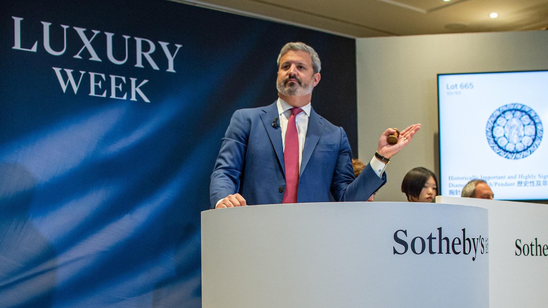 Un uomo in abito blu si trova su un podio con la scritta "Sotheby's" durante un evento d'asta alla Settimana del Lusso. Tiene in mano un martelletto, con l'immagine dell'oggetto e il numero del lotto proiettati sullo sfondo.