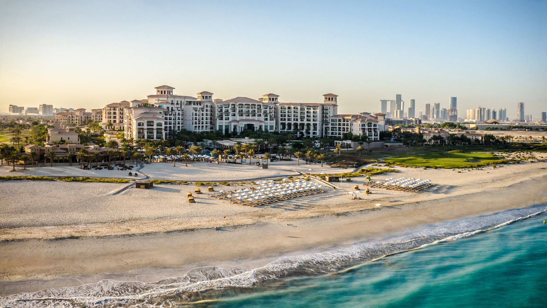 Un grande resort sulla spiaggia con file di ombrelloni e sedie a sdraio sull'arenile, di fronte all'acqua turchese. I moderni grattacieli della città si ergono sullo sfondo lontano, sotto un cielo limpido.