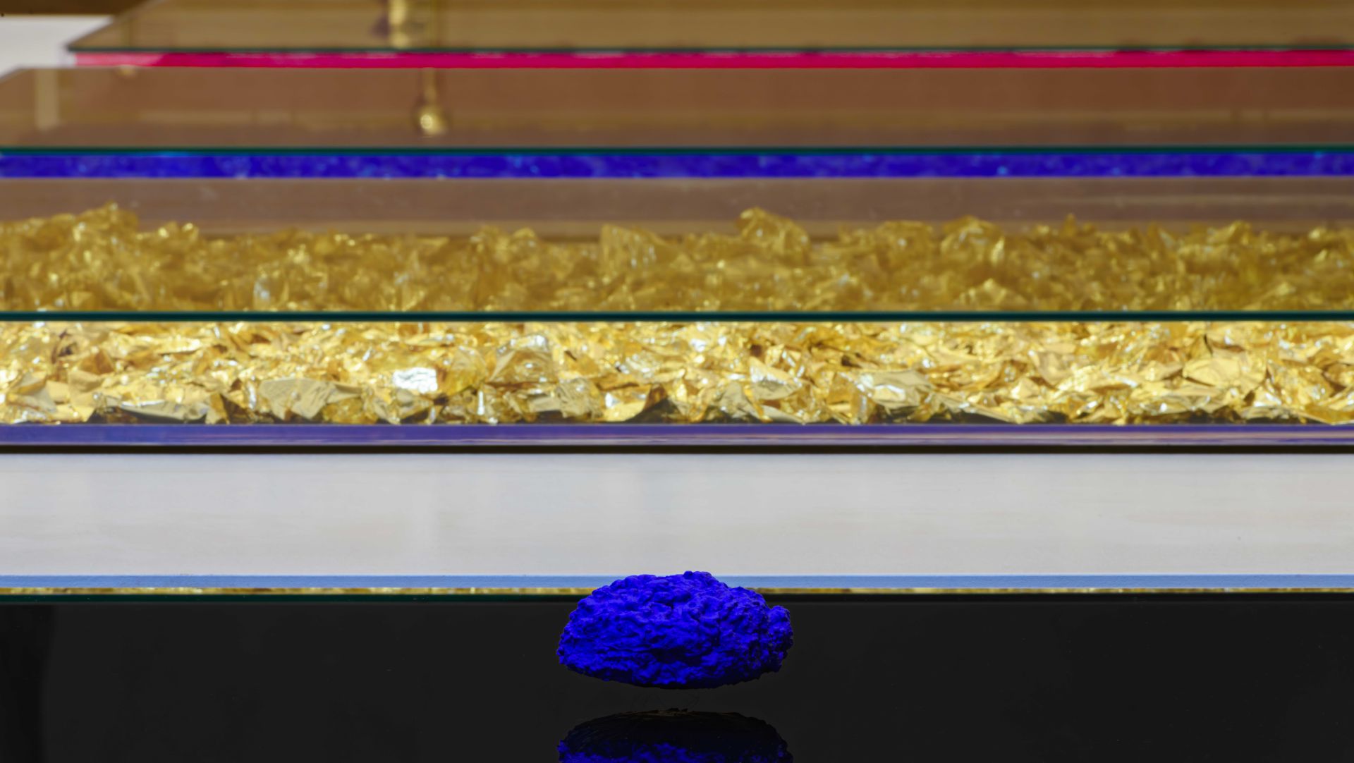 Una roccia blu vivida si trova su una superficie nera riflettente, con sopra file di pannelli di vetro contenenti frammenti d'oro lucido e bande di colori rossi e blu sullo sfondo.