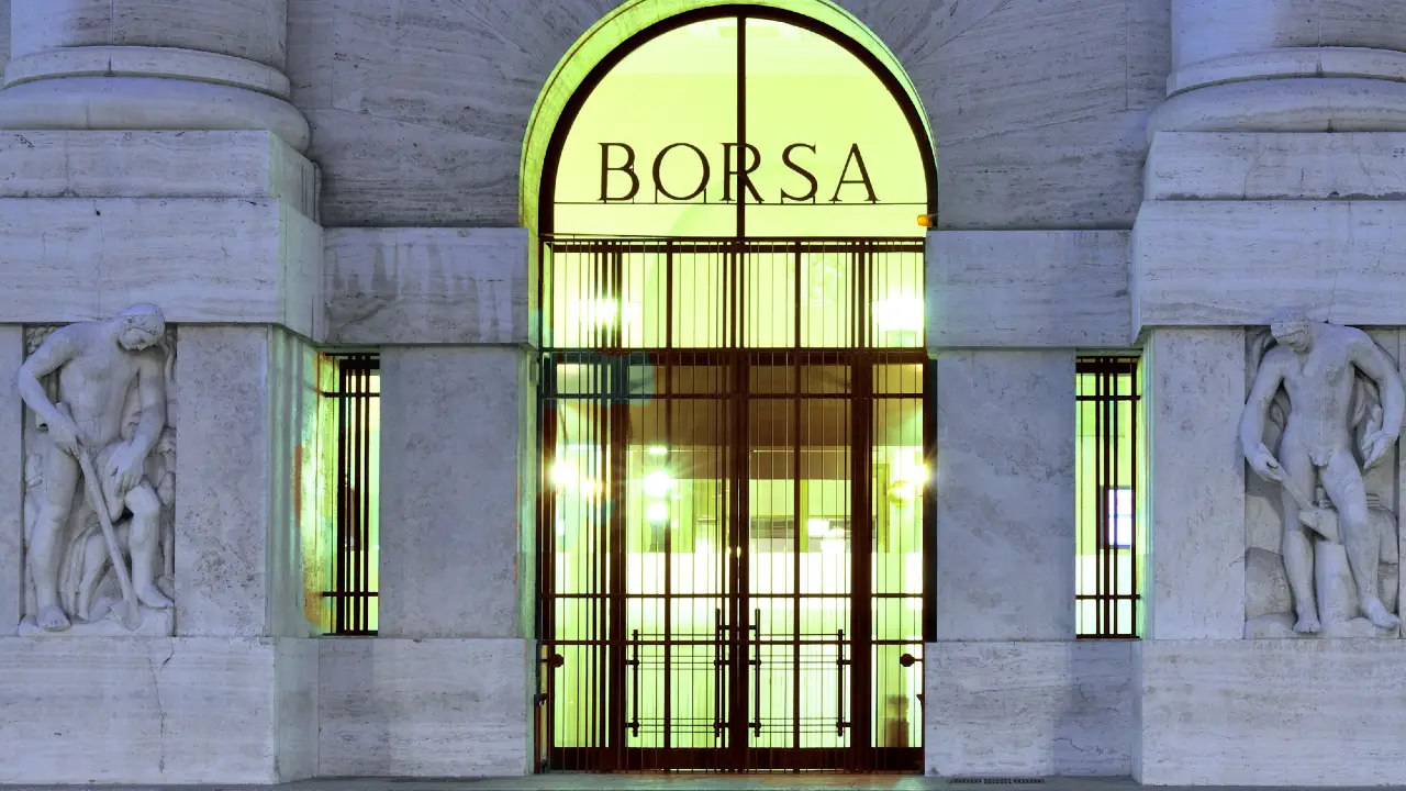 Un'alta porta di vetro con la scritta "BORSA", inserita in un edificio in pietra con statue ai lati e luci brillanti all'interno.
