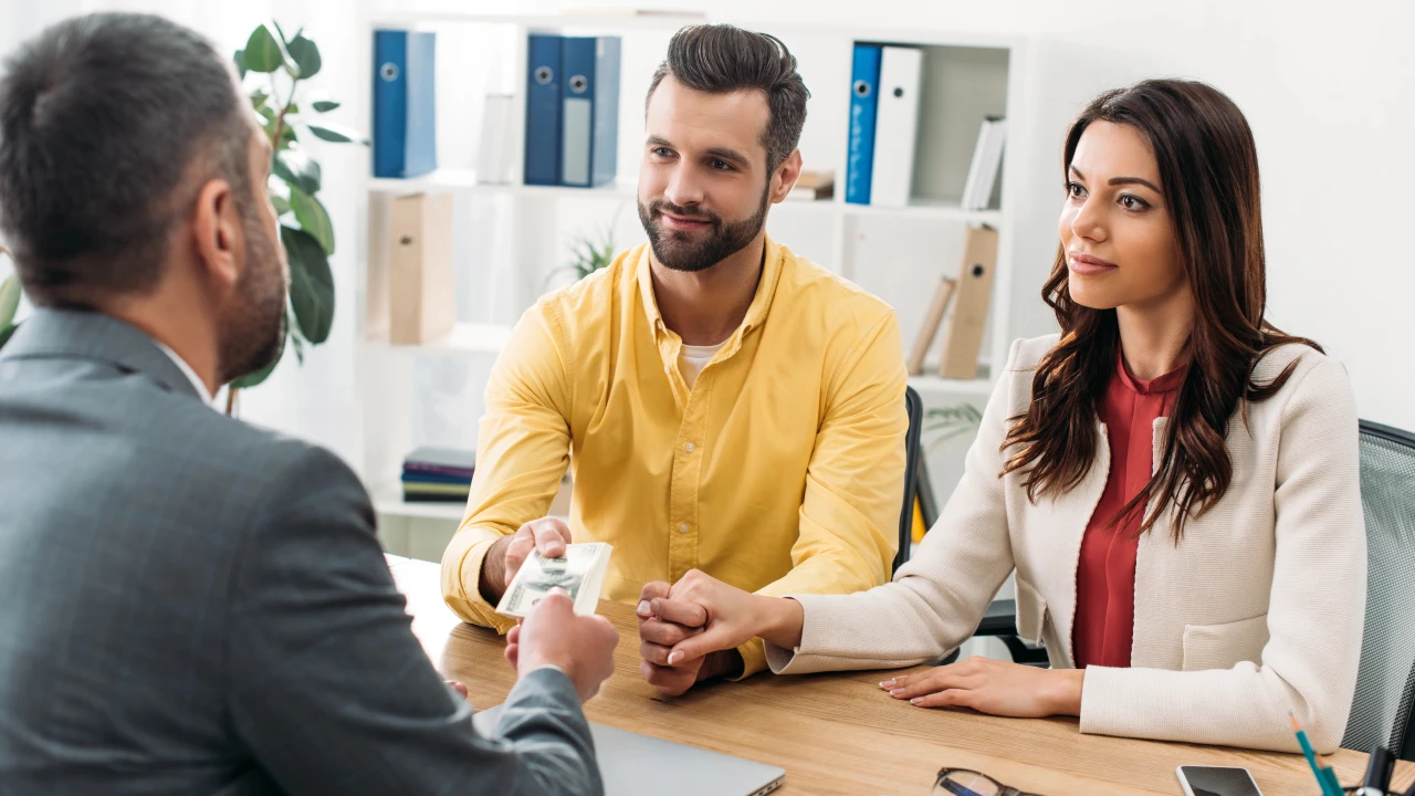 Un uomo e una donna siedono vicini a una scrivania, tenendosi per mano. L'uomo dà dei contanti a un consulente finanziario in giacca e cravatta di fronte a loro. Sullo sfondo sono visibili scaffali e piante dell'ufficio.