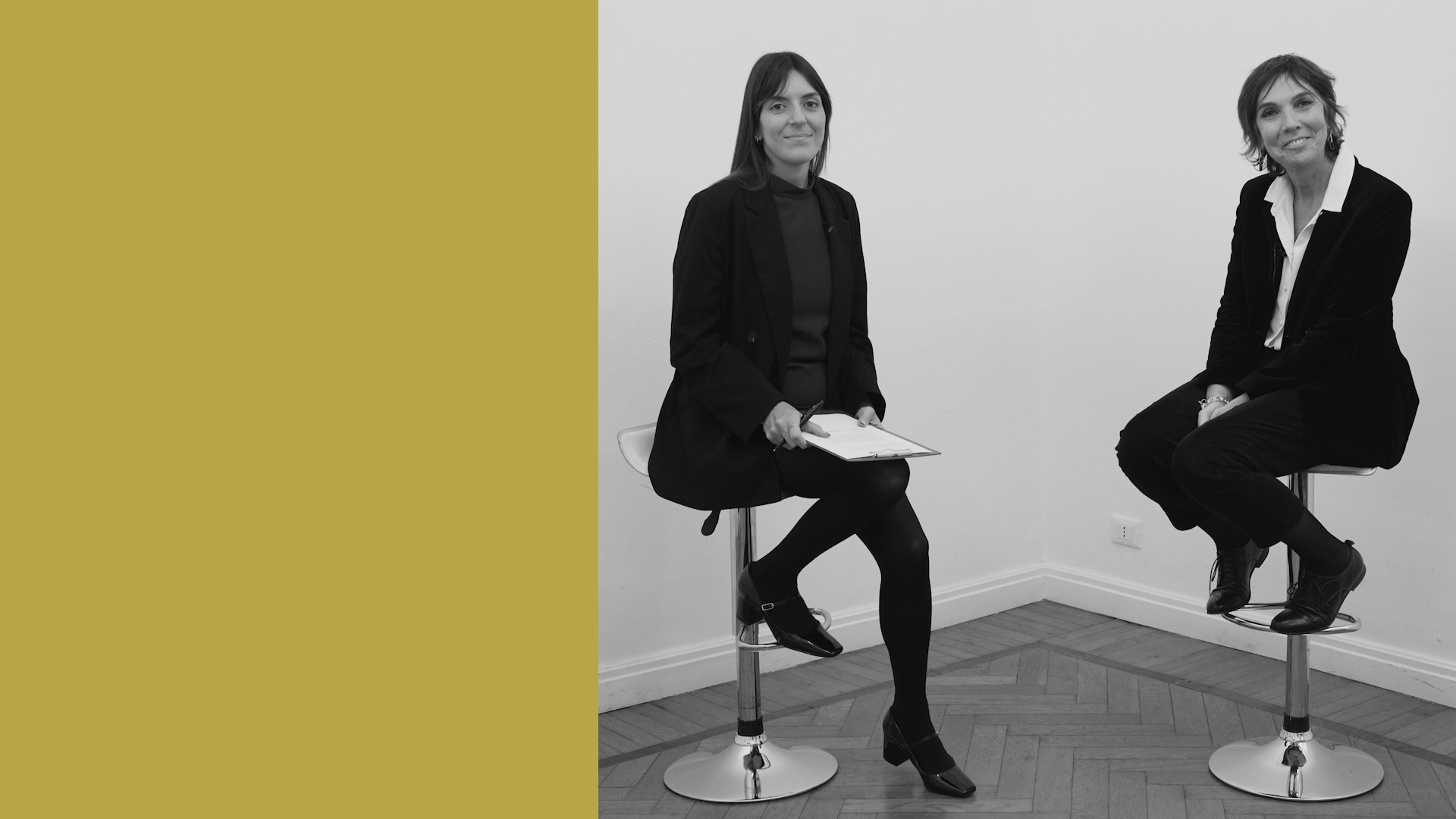 Due donne in abiti da lavoro siedono su sgabelli da bar in un angolo di una stanza con pareti bianche e pavimento in legno. La metà sinistra dell'immagine è di colore giallo senape.