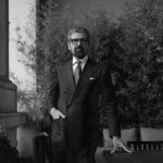 Un uomo in abito scuro e cravatta sta all'aperto accanto a un tavolo, con una mano in tasca e l'altra appoggiata sul tavolo. Sullo sfondo, piante alte e un muro di pietra. L'immagine è in bianco e nero.