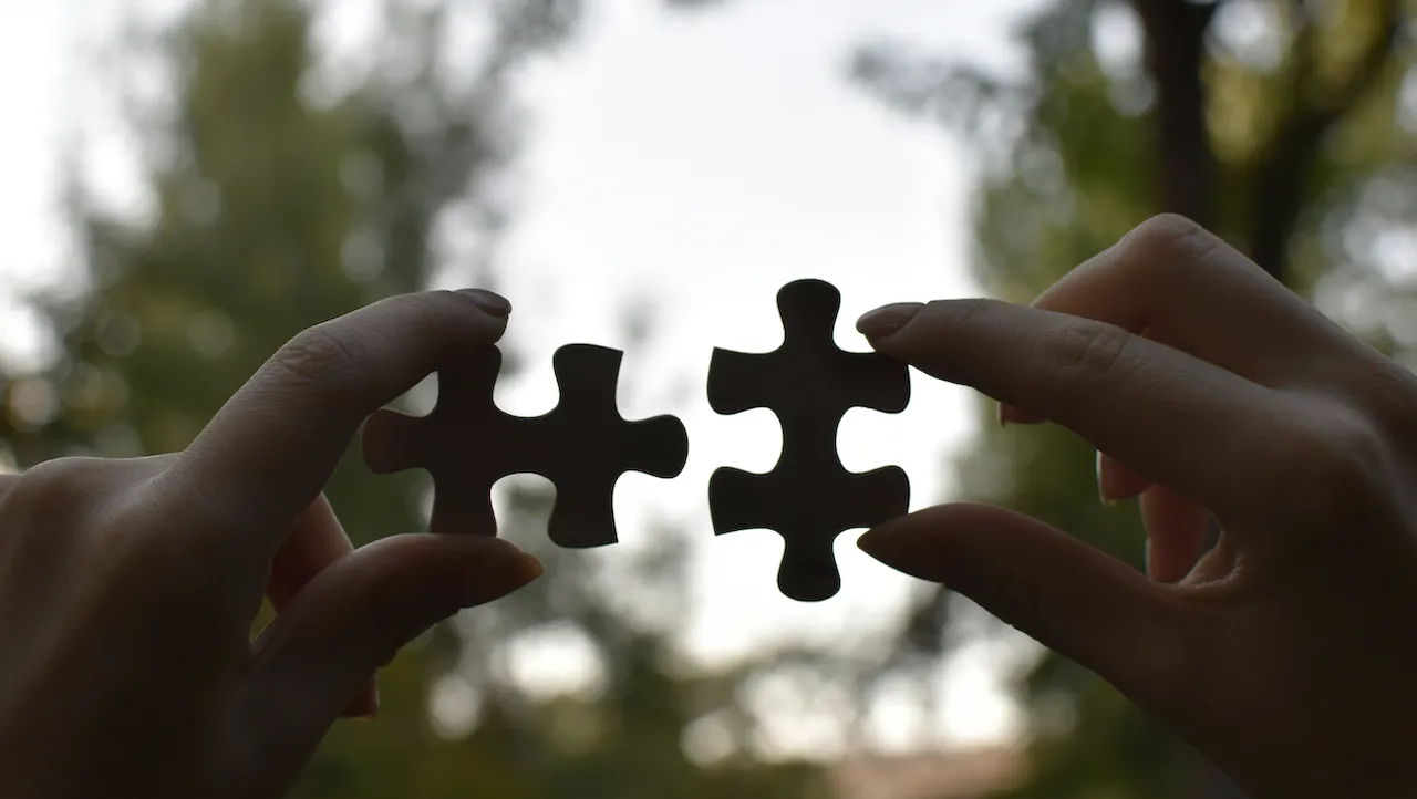Due mani sorreggono pezzi di puzzle separati su uno sfondo esterno sfocato con alberi, con i pezzi posizionati come se stessero per essere uniti.